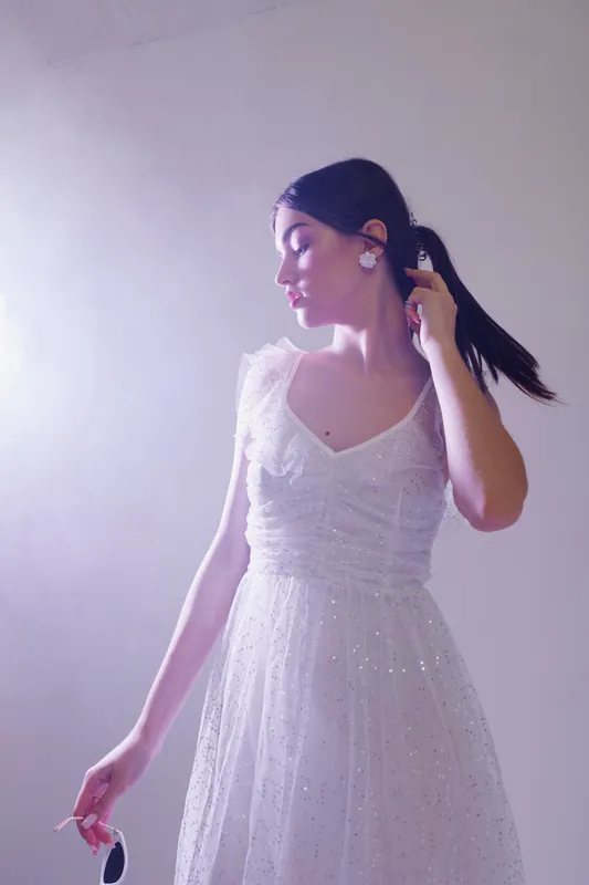 Young woman in a white dress poses elegantly while holding sunglasses. Soft lighting highlights her beauty and fashion style with a calm expression on a light background.