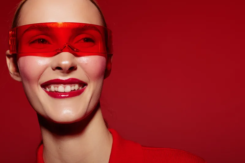 Smiling woman wearing red futuristic visor and red outfit poses against red background. Concept of modern fashion, technology, and vibrant style with bright makeup and confident expression.