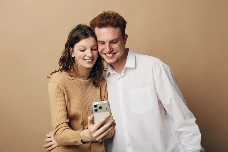 Smiling young couple takes selfie together on smartphone. Woman in brown sweater and man in white shirt pose happily against beige background. Concept of friendship, technology, and casual lifestyle.