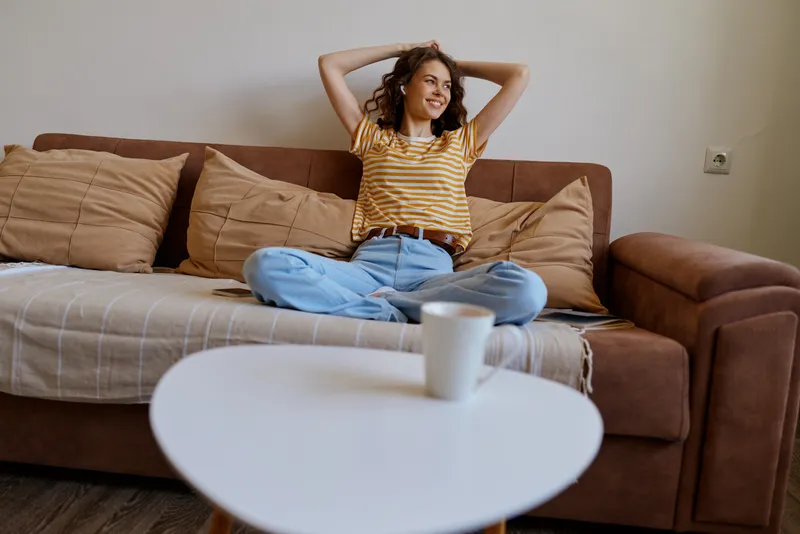 A relaxed woman sits on a brown sofa at home, legs crossed, hands behind her head and a smile. Bright living room ambience conveys comfort, tranquility, and everyday wellness.