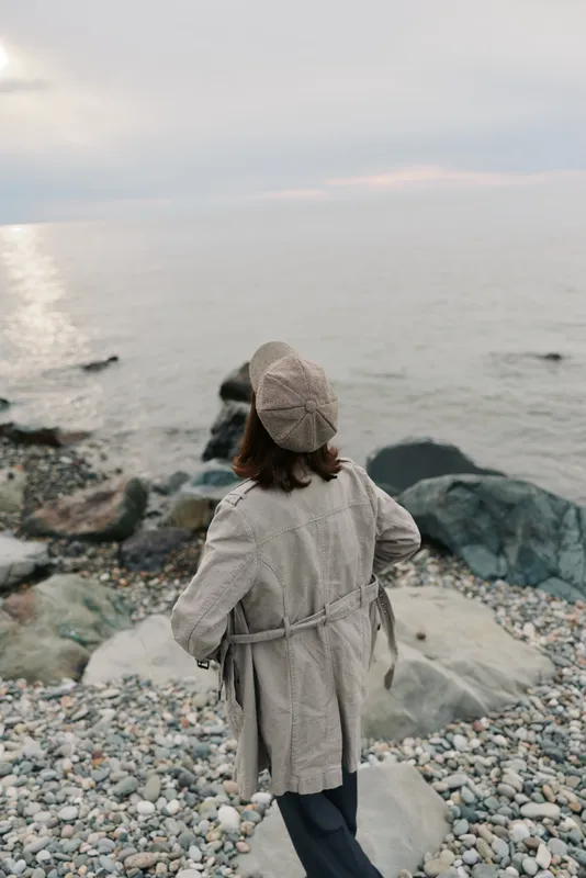 Woman in beige coat and hat stands on rocky beach looking at calm sea under cloudy sky. Concept for autumn, solitude, nature, and peaceful coastal landscape.