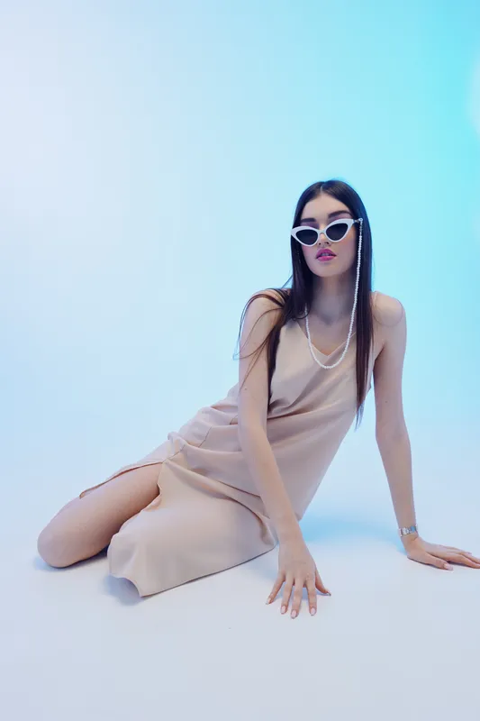 Young woman in a beige dress poses on the floor wearing white sunglasses and layered necklaces. Studio shot with soft lighting on a blue gradient background. Concept of beauty fashion style.