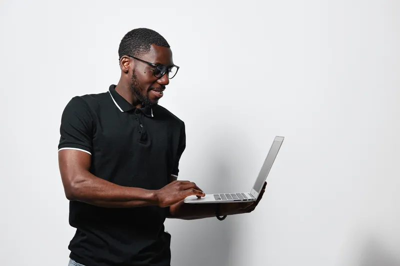 African American man with dark skin wearing glasses and black polo shirt using laptop computer for work or study on white background. Technology, casual and focused.