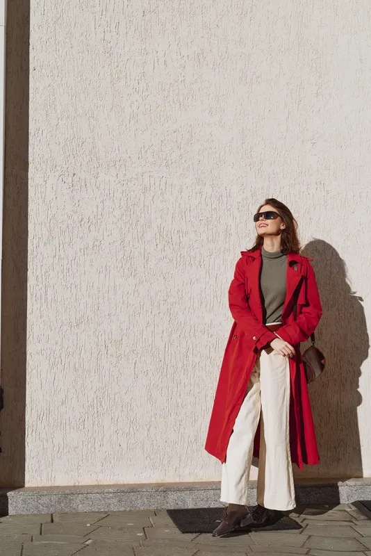 Stylish woman in red coat and sunglasses stands against textured wall in sunlight, wearing white pants and brown shoes, holding small brown purse, casual urban fashion look.