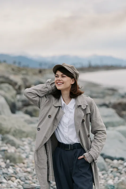 Smiling woman in warm knitted coat and cap stands on rocky beach with hand behind head. Overcast sky and distant mountains create calm natural atmosphere for outdoor fashion concept.
