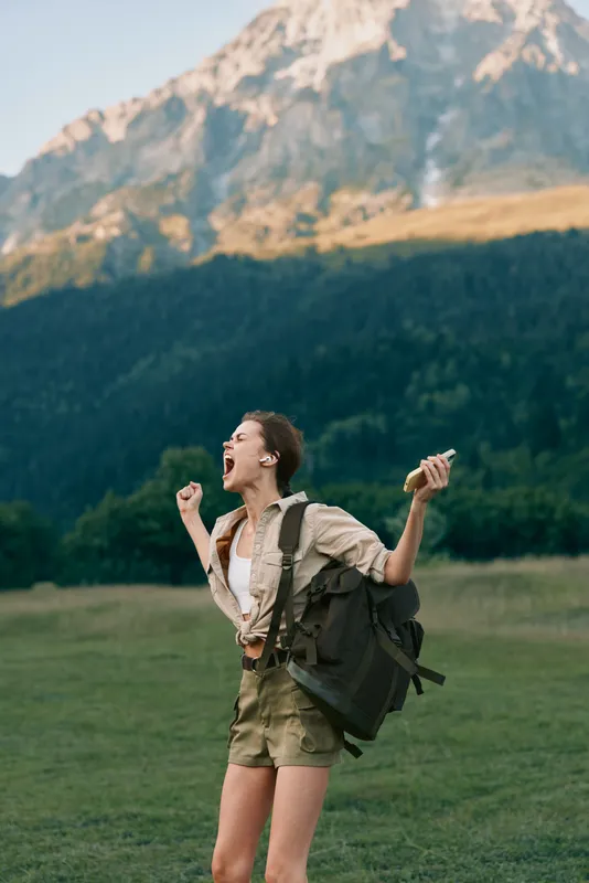 One person, a young woman hikes with a backpack through a vast green meadow, celebrating outdoors with towering mountains rising in the distance and a clear blue sky above.