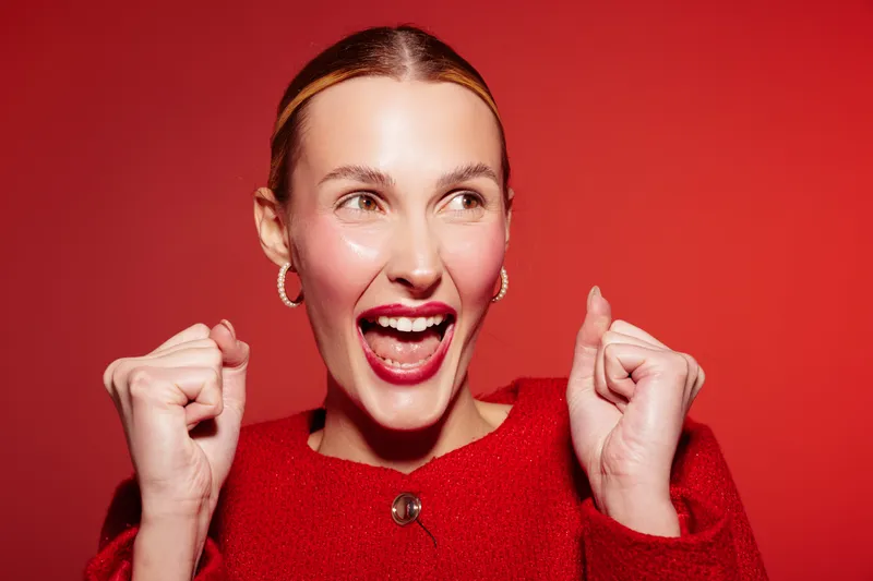 Excited young woman in red sweater smiles widely with raised fists on red background. Concept of happiness, celebration, success, and positive emotions in studio portrait.