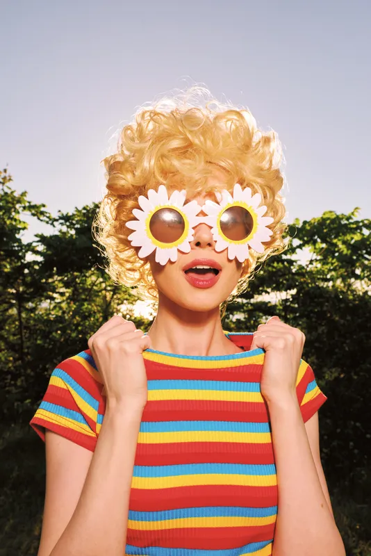 A playful woman in a colorful striped shirt wears oversized flower sunglasses in bright sunlight, conveying retro whimsy and fashion experimentation.