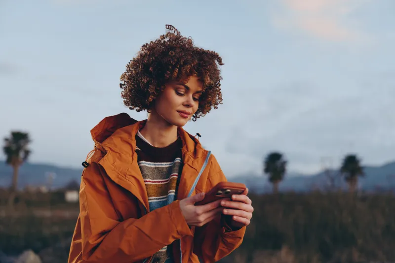 Woman lifestyle outdoors with curly hair and an orange jacket, smiling portrait while holding a smartphone, countryside field and distant mountains under soft evening sky, casual travel leisure
