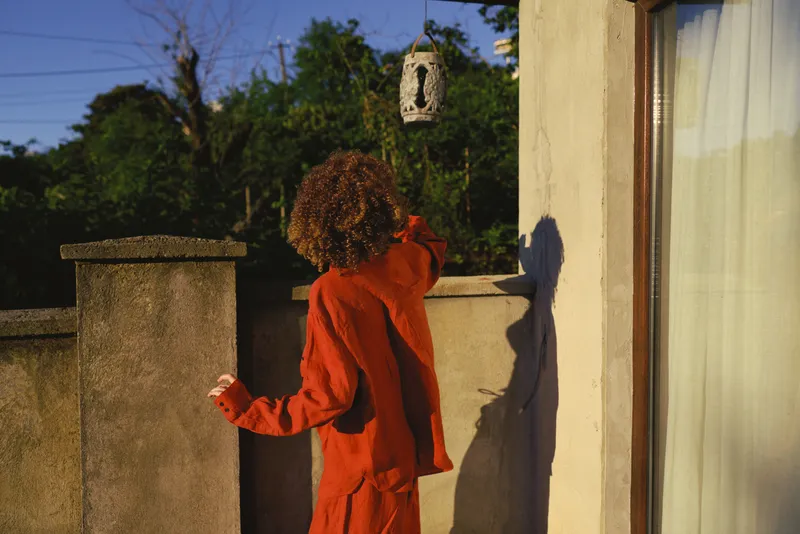 A woman with curly hair in a bright red outfit stands on a sunlit balcony, reaching toward a hanging lantern, capturing a moment of curiosity and quiet exploration