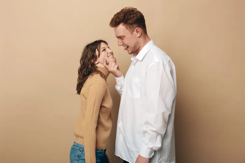 Smiling young couple in casual clothes share a tender moment, man gently touches woman’s chin. Beige background, warm lighting, concept of love, affection, and relationship happiness.