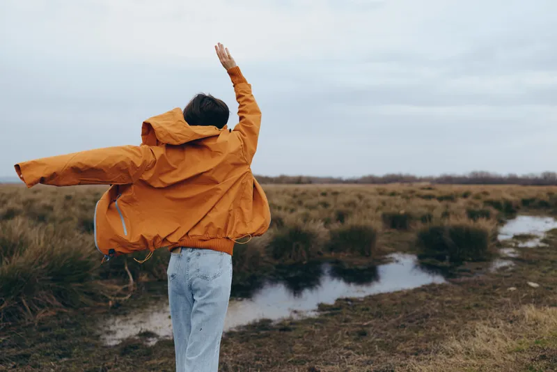 Woman lifestyle outdoors field nature raincoat cloudy sky, orange coat, jumping with arms raised by water in windy grassland, enjoying freedom, casual travel mood, scenic background