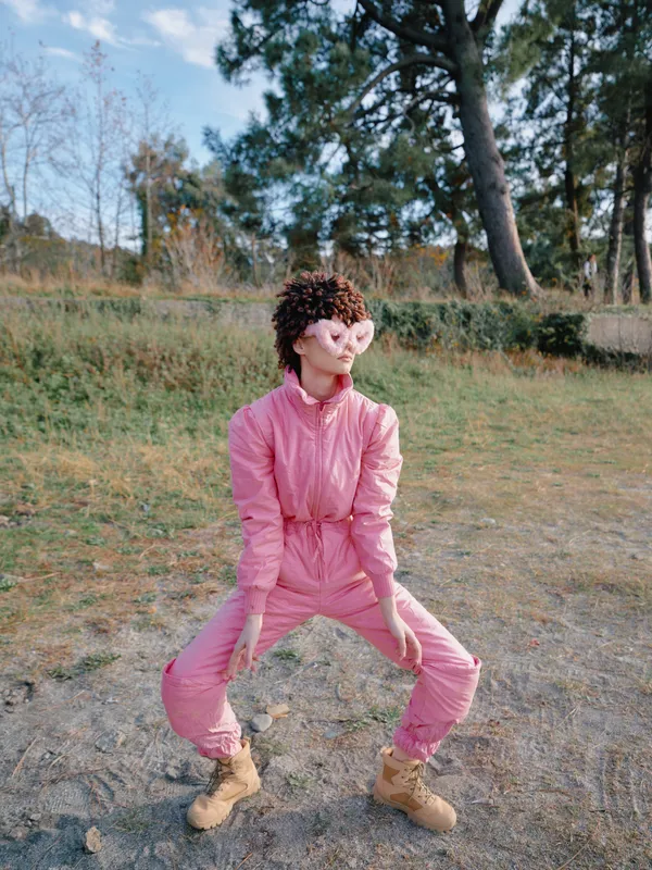 Outdoor person in a pink jumpsuit strikes a playful pose in a park, boots on dirt, trees in the background, capturing candid fashion and bright daytime vibes