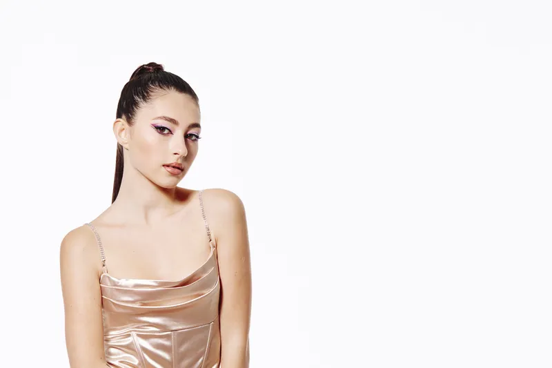 Young woman with creative pink eye makeup poses confidently in a shiny satin dress. Studio shot with minimalistic white background. Concept for beauty fashion and makeup art.