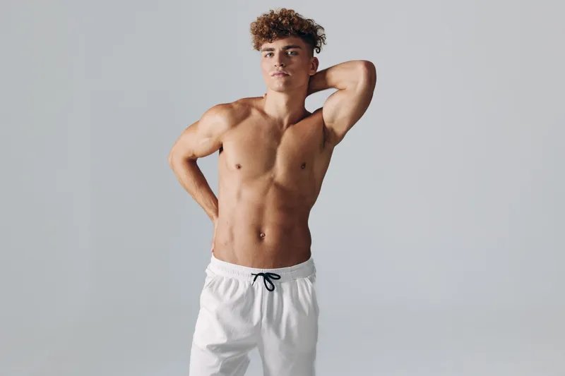 Young man with muscular body posing shirtless in white shorts against gray background. Fitness, health and confident athlete showing toned abs and curly hair in studio lighting.