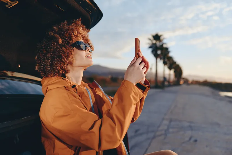 Woman lifestyle outdoors, orange jacket and sunglasses, curly hair in soft wind, standing by a car on an open road at sunset, holding a peace sign, warm natural light, travel vibe