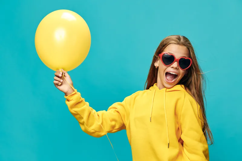 Girl with sunglasses holding yellow balloon and wearing yellow hoodie against blue background. Happy young woman smiling outdoors with vibrant colors and casual outfit.