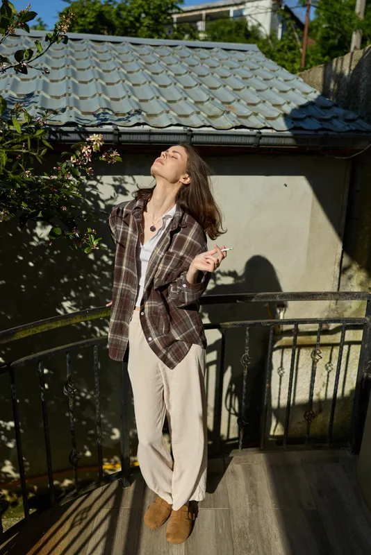 Woman on a balcony posing in a plaid blazer, light shirt, and wide pants under bright sunlight, creating a casual fashion mood with a relaxed stance and warm outdoor atmosphere