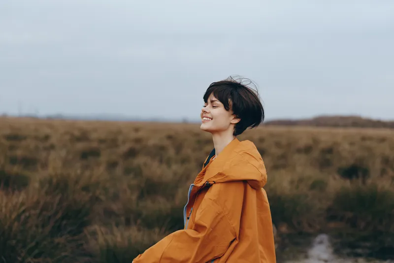 Woman lifestyle outdoors, smiling in profile, wearing orange jacket, standing in grassy field under cloudy sky, soft wind, calm mood, natural colors, distant landscape