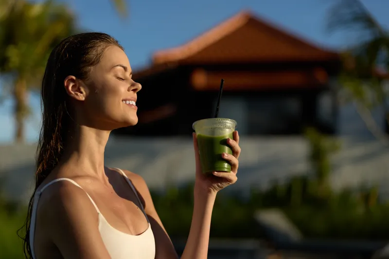 A smiling woman in a white tank top enjoys a green smoothie outdoors, eyes closed and face turned to the sun. The background shows a blurred house and garden. Lifestyle concept.