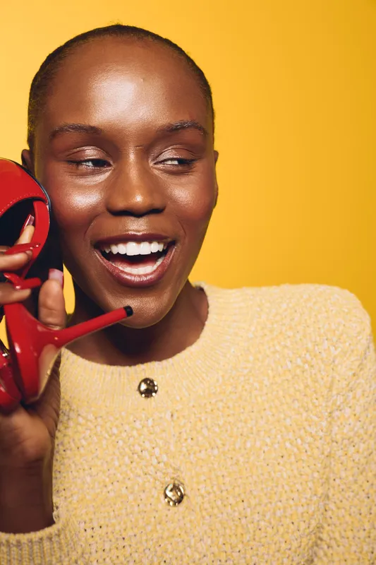 Smiling African woman with dark skin holds red high heel shoe near face, wearing yellow textured sweater. Bright yellow background highlights cheerful mood and fashion concept with beauty focus.