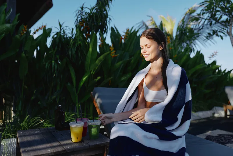 Tropical scene shows a relaxing woman enjoying sunlit moments beside a wooden deck with striped towel and two drinks, conveying calm leisure and warm summer mood.