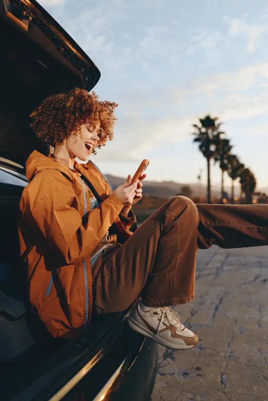 Woman lifestyle outdoors by a car on the road, smiling in warm sunset light, curly hair, jacket and casual pants, peace gesture, natural colors, open sky