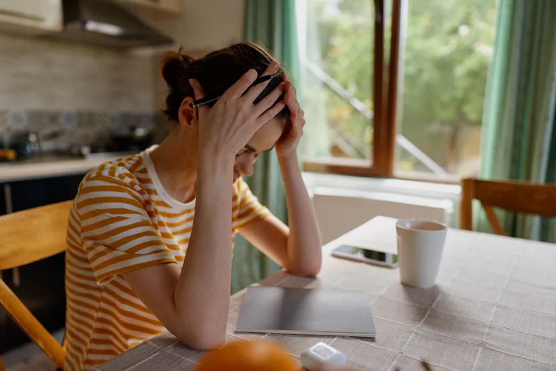 A woman sits at a kitchen table, head in hands, looking stressed and tired. Soft natural light highlights a calm home scene and everyday routine, conveying fatigue and mindfulness.