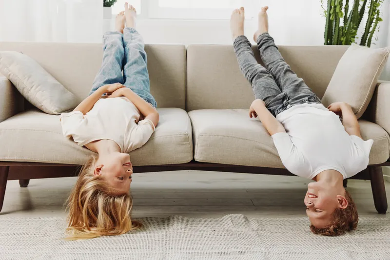 Two happy children lie upside down on a beige sofa in a bright living room. They smile and enjoy playful moments together, creating a joyful and carefree atmosphere.