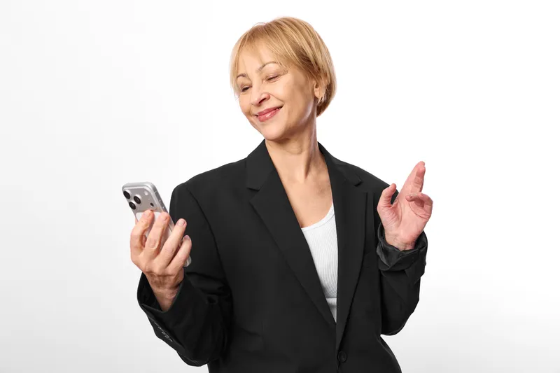 Woman senior in business suit smiles and makes peace gesture, holding smartphone, communication and technology on white studio background, friendly portrait