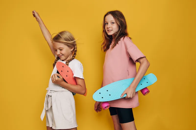 Two happy girls with skateboards stand against yellow background. One girl raises arm while the other smiles holding blue skateboard. Fun active lifestyle concept.
