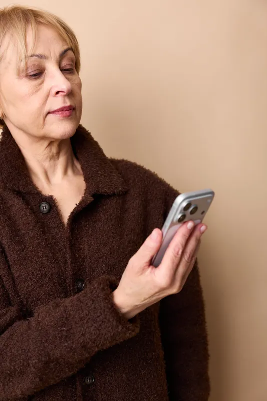 An elderly woman holding a smartphone shows stress and anxiety, eyes closed, conveying loneliness and mental health concerns in quiet indoor communication