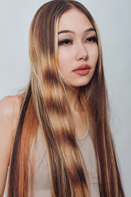 Young woman with long straight hair and natural makeup posing against white background. Portrait of female with relaxed expression and casual sleeveless top in studio light.