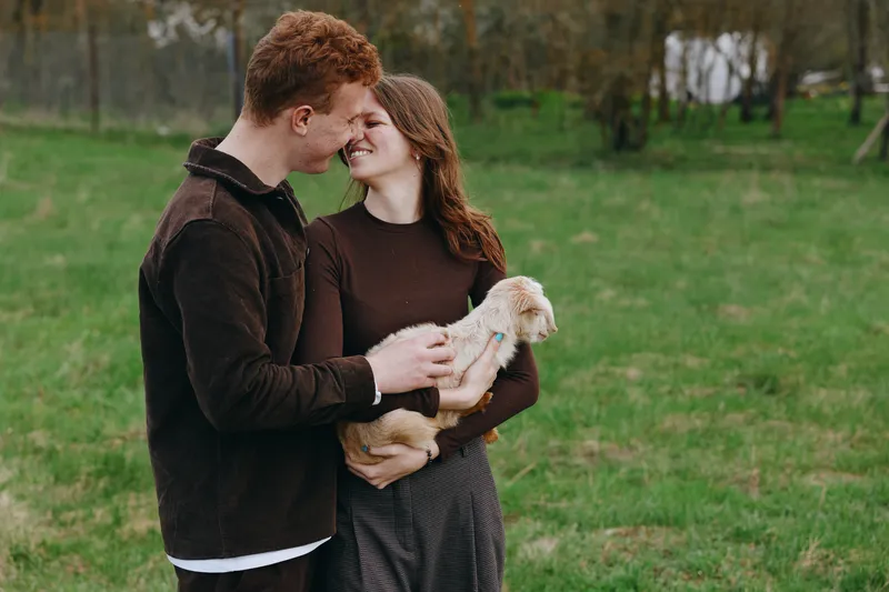 Young couple embraces tenderly outdoors while holding a small baby goat in their arms, sharing a joyful and intimate moment in a natural green field.