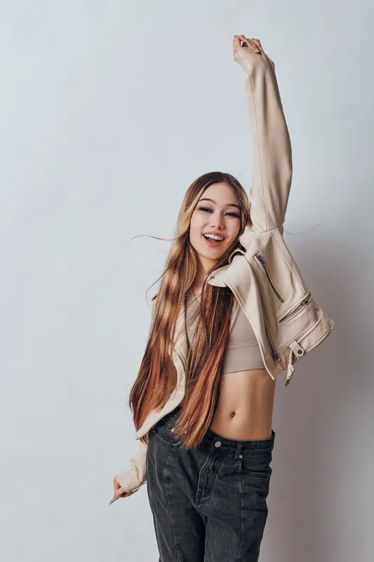 Woman with long hair wearing a crop top and jacket smiling and raising one arm for joy with casual fashion style on a white plain background indoors.