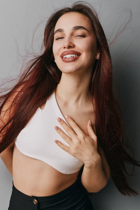Woman with long hair smiling and eyes closed posing in white crop top and black pants against gray background. Portrait of happy female with natural makeup and hand on chest.