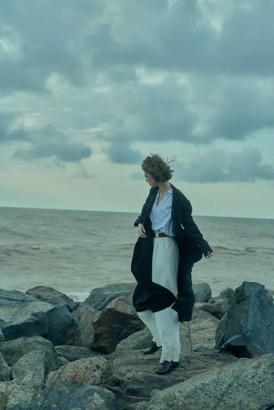 Woman on a rocky shore by the sea, wearing a long coat over a white dress, hair blown by wind, moody clouds above, calm horizon, fashion meets rugged coastscape