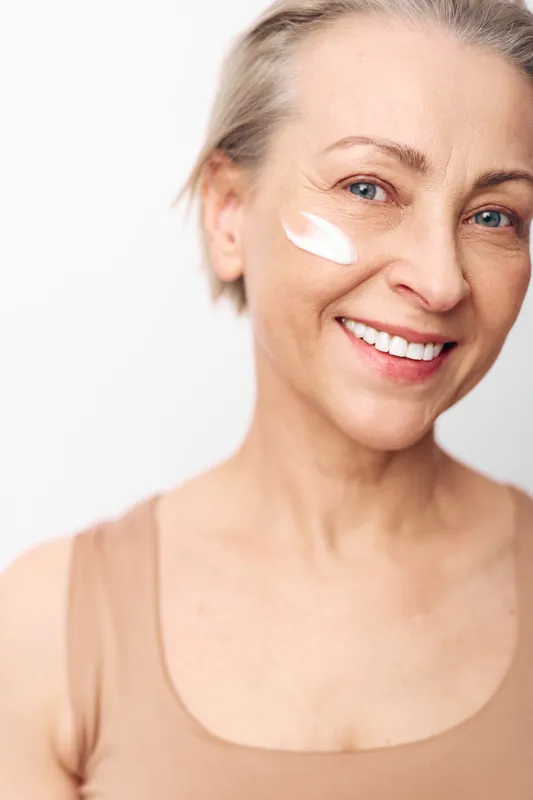Smiling mature woman with blue eyes applies white face cream on cheek in close-up portrait on white background. Concept of skincare, natural beauty, wellness, anti-aging care, and self-care.