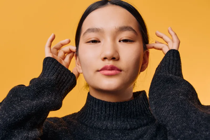Woman with closed eyes and relaxed face touching ears wearing dark sweater with turtleneck posing on yellow background for portrait and beauty concept closeup.