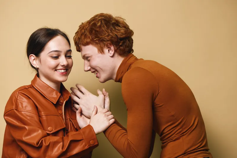 Young man and woman smiling and holding hands, wearing casual autumn outfits with tender emotion and friendly connection in studio with beige background.