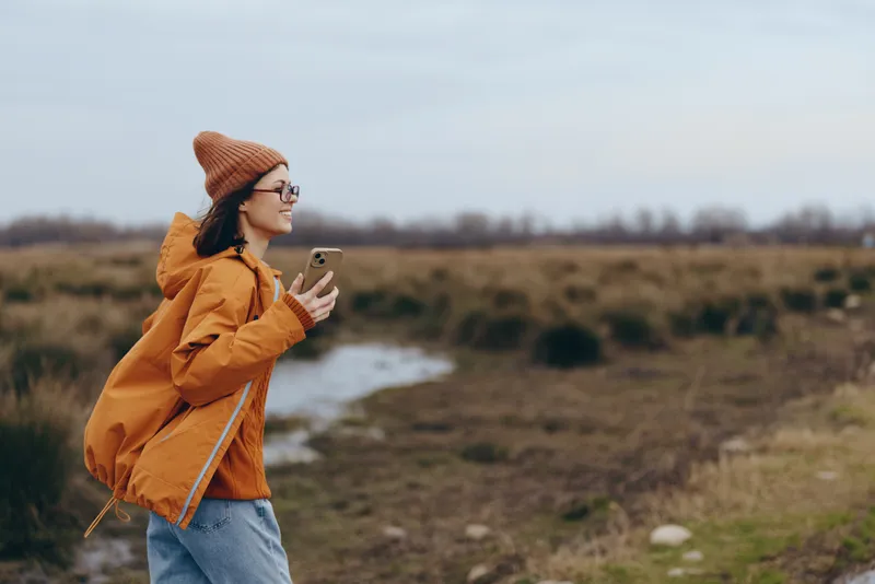 Woman lifestyle outdoors in rural field, orange jacket and knit hat, holding smartphone to take a photo, portrait with nature scenery, calm countryside and autumn background, smile walking