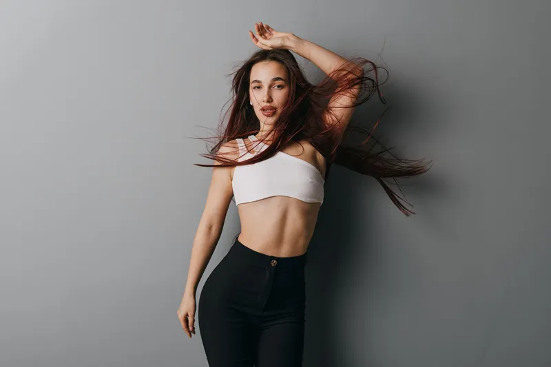 Young woman posing with flowing hair wearing white crop top and black high waist pants against grey background for fashion and lifestyle portrait.