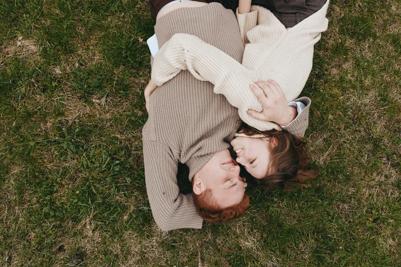 Candid couple outdoors on grass, embracing, wearing sweaters, sunlight highlights their connection, calm mood, natural setting, warm tones, relaxed lifestyle, intimate moment