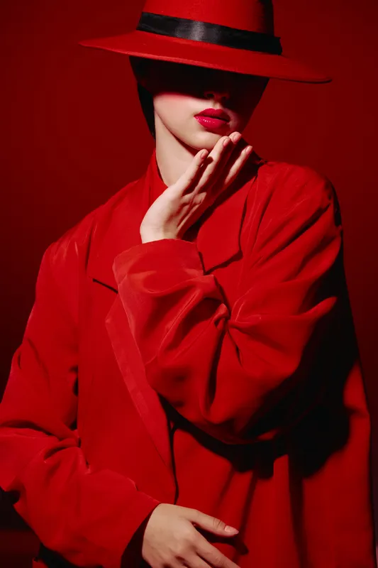 Stylish woman wearing a red coat and matching hat with bold red lipstick touches her face against a vivid red background. Creative beauty fashion concept.