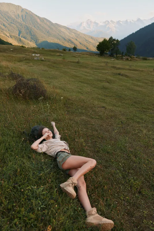 Young woman rests in a sunlit meadow with distant mountains, barefoot, relaxed and smiling softly at the serene landscape.