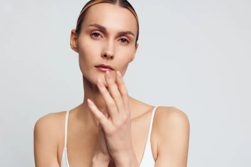 Serious woman with natural makeup looking at camera, soft lighting, neutral background, minimalistic style, calm expression, bare shoulders visible