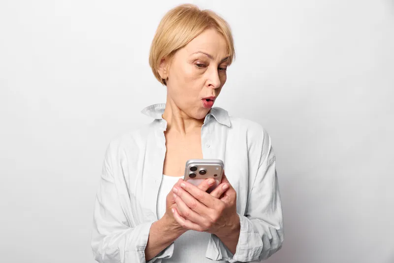 Surprised woman holding smartphone with worried face, reading text on screen, isolated on white background, close up portrait, mobile technology communication