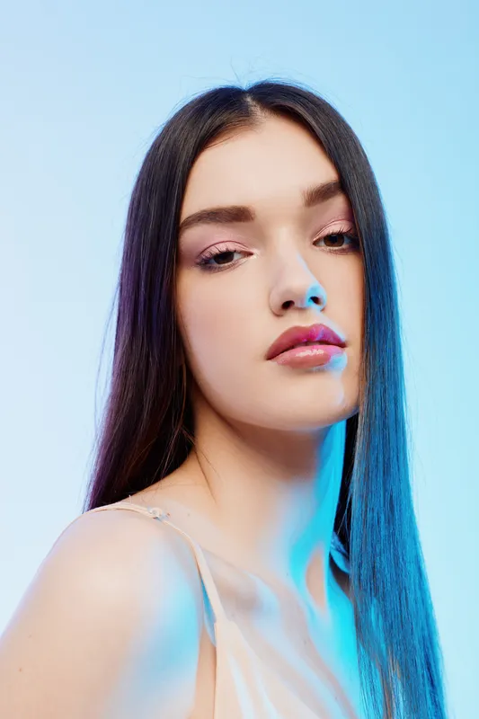 Portrait of a woman beauty fashion model with long straight hair and natural makeup. Soft lighting highlights her flawless skin on a blue background. Concept of elegance and style.