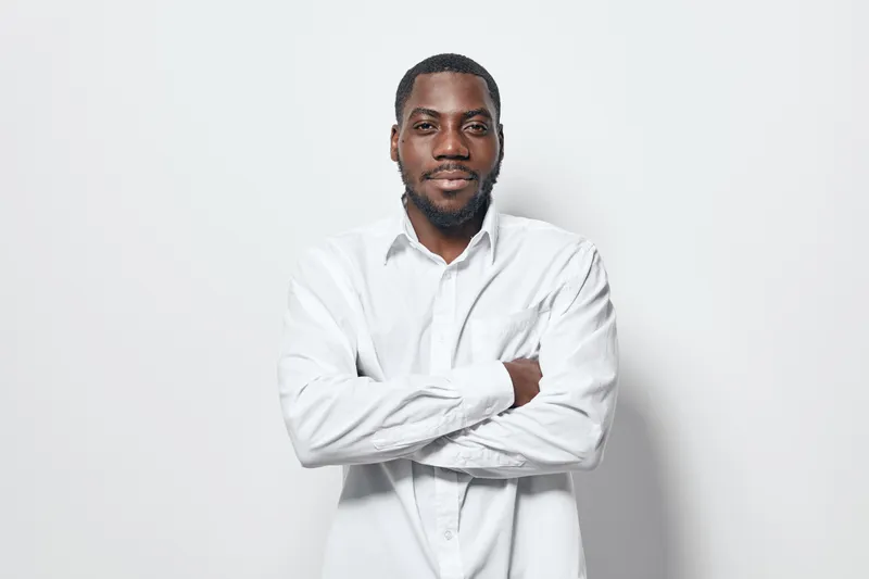 African American man with dark skin wearing white shirt standing with arms crossed against white background looking confident and relaxed in studio portrait.