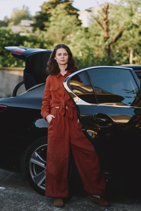 Confident woman in a burnt orange jumpsuit stands beside a sleek car in an outdoor setting. Natural daylight highlights texture and style, conveying modern fashion and vibe